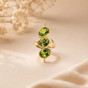 Könnte beinhalten: Ein Goldring mit drei vertikal angeordneten, oval geschliffenen grünen Peridot-Edelsteinen, die jeweils mit einem winzigen eingebetteten rosa Stein akzentuiert sind. Die Ringschiene besteht aus poliertem Gelbgold mit klarem Design.