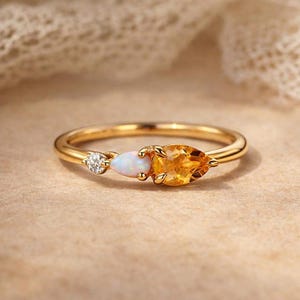 Könnte beinhalten: Ein Goldring mit drei Steinen: einem kleinen runden klaren Edelstein, einem birnenförmigen weißen Opal und einem größeren birnenförmigen gelben Citrin. Die Steine sind in eine dünne, polierte Goldschiene mit filigranem Design gefasst.