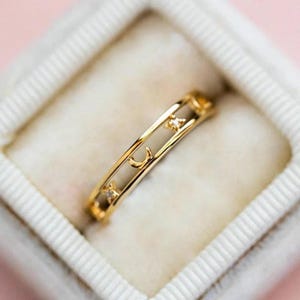 Könnte beinhalten: Ein goldfarbener Ring mit einem Himmelsdesign mit Sternen und einem Mond. Der Ring ist mit kleinen, klaren Edelsteinen besetzt und wird in einer weißen Ringschatulle präsentiert.