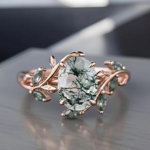Könnte beinhalten: Roségoldener Ring mit einem ovalen Moosachat-Edelstein. Der Ring hat ein von der Natur inspiriertes Design mit Blatt- und kleinen Edelsteinakzenten. Der Moosachat ist hellgrün mit dunkelgrünen Einschlüssen.