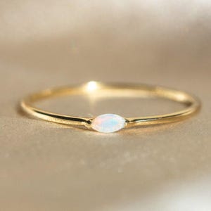 Könnte beinhalten: Ein zarter Goldring mit einem opalartigen Edelstein in Marquiseform. Der Ring hat ein dünnes, poliertes Band und einen schimmernden, irisierenden Opal. Der Ring ist vor einem weichen, neutralen Hintergrund platziert.
