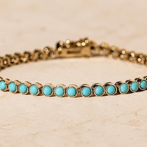 Könnte beinhalten: Ein goldfarbenes Tennisarmband mit einer durchgehenden Reihe kleiner, runder, leuchtend türkisfarbener Steine in einzelnen Metallfassungen, die durch bewegliche Glieder verbunden und mit einem rechteckigen Kastenschloss gesichert sind.