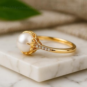 Könnte beinhalten: Ein goldener Ring mit einer großen, glänzenden weißen Perle. Der Ring ist mit kleinen, funkelnden Edelsteinen besetzt. Der Ring liegt auf einer weißen Marmoroberfläche. Der Text "Shubhkrishnajewells" ist sichtbar.