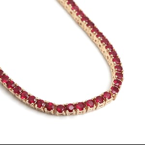 Könnte beinhalten: Eine goldfarbene Halskette mit einer Reihe leuchtend roter Edelsteine. Die Edelsteine sind gleichmäßig angeordnet und in einem goldenen Metall gefasst. Die Halskette wird vor einem weißen Hintergrund gezeigt, um das Design des Schmucks hervorzuheben.