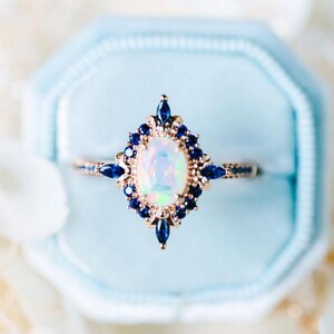 May include: A rose gold ring with an oval opal center stone, surrounded by small, dark blue gemstones. The ring is set in a light blue ring box. The design features a starburst pattern around the opal, with additional blue stones along the band.