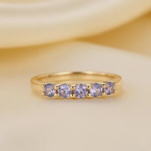 Könnte beinhalten: Ein goldfarbener Ring mit fünf runden, hellvioletten Edelsteinen in einer Reihe. Der Ring befindet sich auf einem weichen, hellgelben Hintergrund. Die Edelsteine sind gleichmäßig angeordnet.