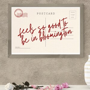 May include: A framed postcard print with the words "feels so good to be in bloomington" in red script. The postcard has a vintage design with a post office stamp and lines for the address. A white vase and flowers are in the foreground.
