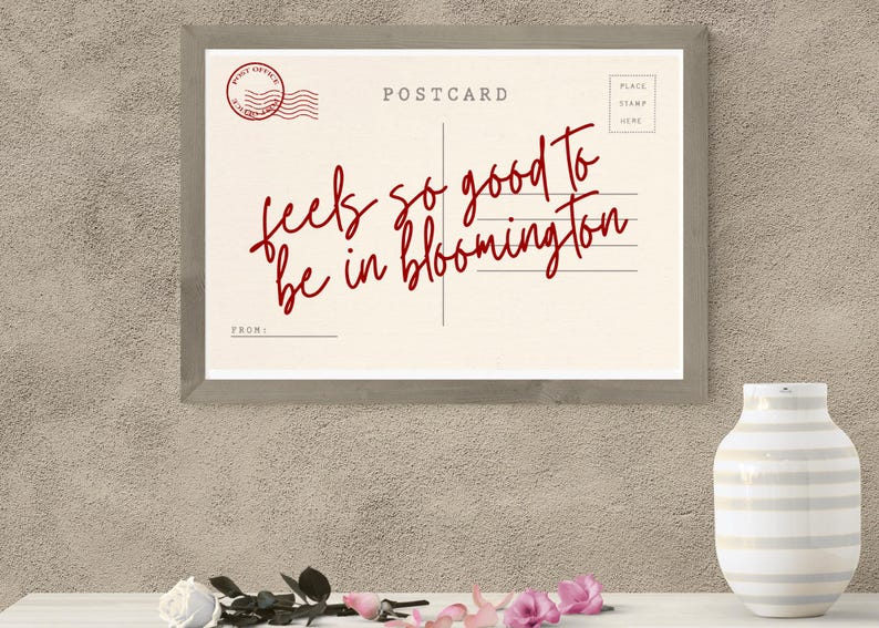 May include: A framed postcard print with the words "feels so good to be in bloomington" in red script. The postcard has a vintage design with a post office stamp and lines for the address. A white vase and flowers are in the foreground.