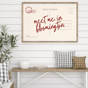 May include: A framed postcard print with the text "meet me in bloomington" in red script. The print is displayed above a wooden console table with decorative pillows and a plant.