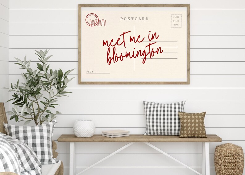 May include: A framed postcard print with the text "meet me in bloomington" in red script. The print is displayed above a wooden console table with decorative pillows and a plant.