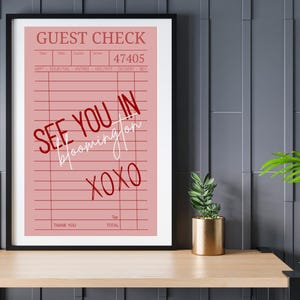May include: A framed pink guest check print with the words "SEE YOU IN Bloomington XOXO" in red and white script. The print is displayed on a wooden shelf with a small plant in a gold pot.