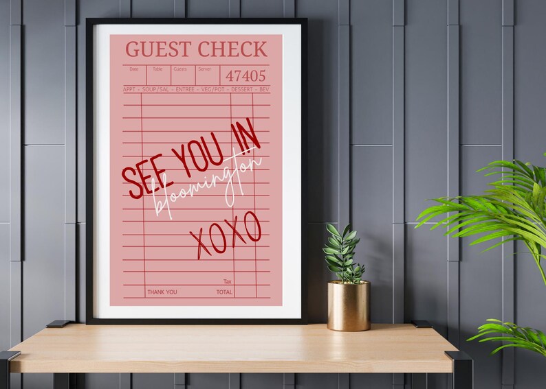 May include: A framed pink guest check print with the words "SEE YOU IN Bloomington XOXO" in red and white script. The print is displayed on a wooden shelf with a small plant in a gold pot.