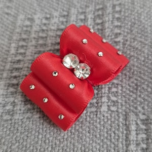 May include: A small, red satin bow with a central cluster of two large, clear rhinestones and several smaller rhinestones. The bow is set against a textured, gray background.