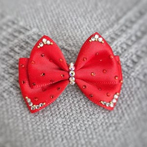 May include: A vibrant red bow with a central rhinestone embellishment and smaller rhinestones outlining the edges. The bow is made of a textured fabric and is set against a soft gray background.