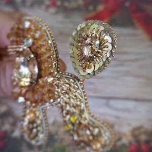 May include: A detailed, handmade brooch in shades of gold and amber. The brooch features a large, teardrop-shaped crystal and is embellished with sequins, beads, and intricate embroidery. The design is ornate and elegant, suitable for special occasions.