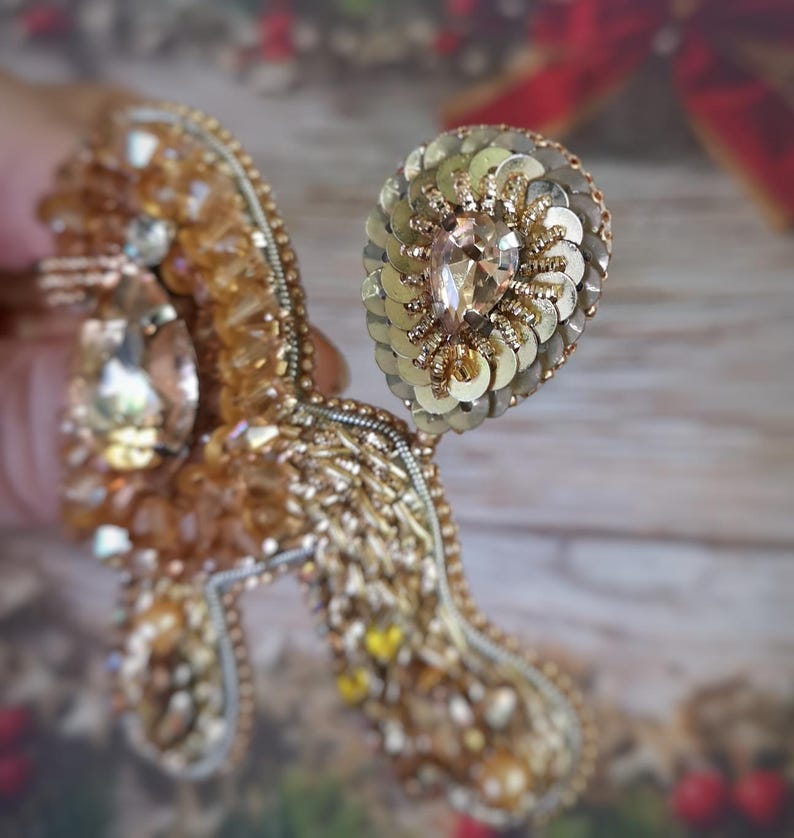 May include: A detailed, handmade brooch in shades of gold and amber. The brooch features a large, teardrop-shaped crystal and is embellished with sequins, beads, and intricate embroidery. The design is ornate and elegant, suitable for special occasions.