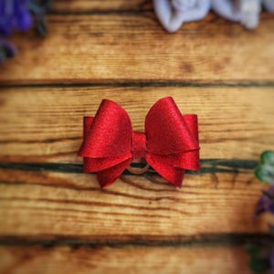 May include: A red glitter bow, likely for hair accessories, is centered on a wooden surface. The bow has a layered design and a small elastic band. The background is a warm, wooden texture with blurred floral accents.