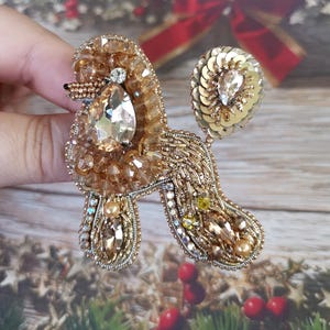 May include: A gold-toned poodle brooch, intricately beaded with crystals and sequins. The brooch features a large, teardrop-shaped crystal at the center, with smaller crystals and beads outlining the poodle's form. The tail is a cluster of gold sequins.