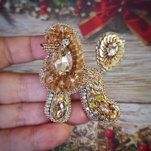 May include: A handcrafted, gold-toned poodle brooch. The brooch is embellished with beads, sequins, and crystals. The poodle's body features a large, teardrop-shaped crystal. The brooch is a unique accessory.