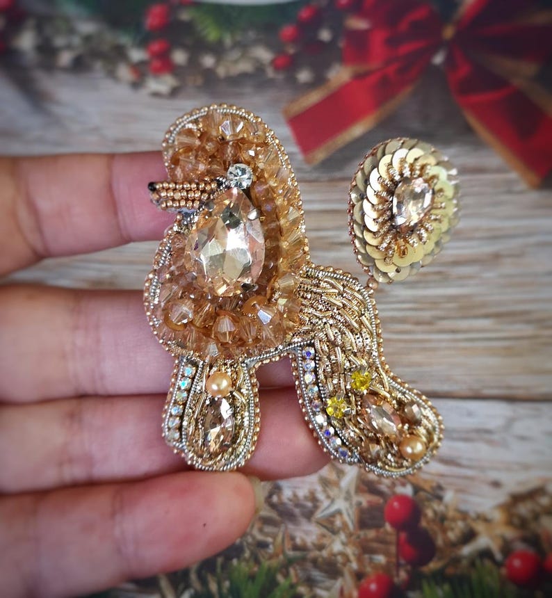 May include: A handcrafted, gold-toned poodle brooch. The brooch is embellished with beads, sequins, and crystals. The poodle's body features a large, teardrop-shaped crystal. The brooch is a unique accessory.