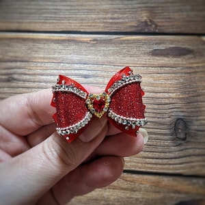 May include: A red glitter bow with a heart-shaped jewel in the center. The bow is adorned with silver rhinestones along the edges. The bow is approximately 3 inches wide.
