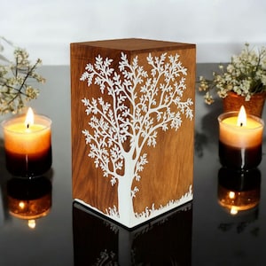 May include: A wooden box featuring a white tree design, with lit candles and flowers. The box is brown with a white tree cut-out. The candles have a colour gradient, and the flowers are white.