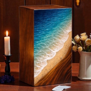 Könnte beinhalten: Holzurne mit Strandmotiv. Die Vorderseite zeigt einen blauen Ozean mit weißen Wellen, die auf einen sandigen Strand treffen. Zwei brennende Kerzen und eine Blumenvase stehen auf dem Tisch.