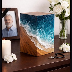 May include: A wooden cremation urn with a unique design. The front of the urn features a detailed ocean wave design with blue and white resin over a wooden base. The top is a solid wood. A memorial photo and flowers are in the background.