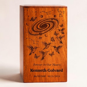 Puede incluir: Urna de madera rectangular con un grabado detallado de una galaxia, constelaciones, colibríes y mariposas. El texto "Forever in Our Hearts", un nombre y fechas también están grabados. La urna es de madera de tono cálido.