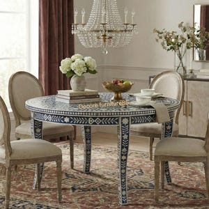 Puede incluir: Un juego de comedor con una mesa redonda con un diseño floral azul marino y blanco, rodeada de cuatro sillas tapizadas beige. Una lámpara de araña cuelga sobre la mesa, y un aparador y un espejo están en el fondo. La habitación tiene una alfombra y cortinas burdeos.