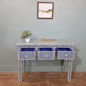 May include: A blue and white patterned console table with three drawers, each with a blue interior. The table has a matching pattern on the legs and top. A small potted plant and artwork are visible.