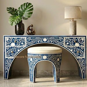 Könnte beinhalten: Ein marineblauer Schminktisch und ein passender Hocker mit weißen floralen Intarsien. Der Tisch hat zwei Schubladen auf jeder Seite und einen geschwungenen Bogen für den Hocker. Eine Lampe und Dekorationsgegenstände stehen oben.