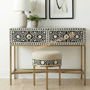 May include: A black and white floral patterned console table with two drawers, a matching stool, and a gold metal frame. The table and stool are placed in front of a white wall with framed artwork.