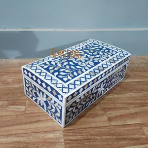 May include: A rectangular blue decorative box with a white floral and geometric inlay design. The box has a hinged lid and is made of a wood-like material. The box is placed on a wooden surface.