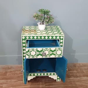 May include: A white and green patterned nightstand with a drawer and cabinet doors. The drawer and cabinet interior are painted blue. A small potted plant sits on top. The nightstand has black and white knobs.