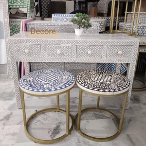 May include: A gray and white bone inlay console table with four drawers and matching round stools. The stools have gold metal frames and patterned tops in blue and white, and black and white. The word "Decore" is visible on the table.