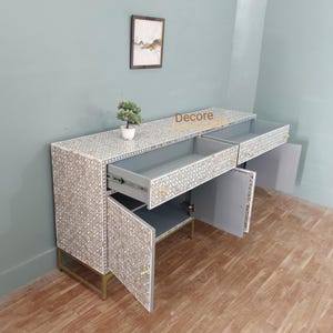 May include: A white and gray patterned cabinet with open drawers and doors. The cabinet has a geometric design and gold-colored legs. A small potted plant sits on top. The word "Decore" is printed on the top of the cabinet.