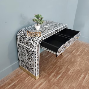 May include: A black and white patterned desk with an open drawer. The desk has a curved design and is decorated with a floral pattern. The drawer has a gold-coloured handle. A small potted plant sits on top of the desk.