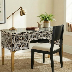 Könnte beinhalten: Ein schwarz-weiß floral gemusterter Schreibtisch mit goldenen Beinen, ein schwarzer Stuhl und eine Messing-Schreibtischlampe. Der Schreibtisch hat mehrere Schubladen und eine zentrale Öffnung. Ein Farn in einem Topf und ein gerahmter botanischer Druck sind ebenfalls zu sehen.