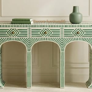 May include: A green and white patterned console table with three drawers and arched legs. The table has brass hardware and is topped with a green vase and two green books. The table is made of wood.