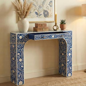 May include: A blue and white floral patterned console table with a drawer open. The table has an arched design and is decorated with a floral inlay. A vase with dried flowers, two wooden boxes, a candle, and a small potted plant are on top.