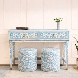 Könnte beinhalten: Ein hellblauer Schminktisch und zwei passende Hocker mit weißem Blumeneinlegearbeiten. Der Tisch hat zwei Schubladen mit goldfarbener Hardware. Die Hocker sind rund und haben das gleiche Blumenmuster. Eine kleine Vase mit einer Pflanze steht auf dem Tisch.