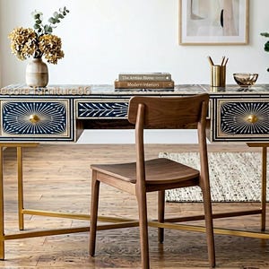 May include: A rectangular desk featuring a dark blue and white patterned inlay on the front drawers and top surface, supported by a gold-toned metal frame. A wooden chair with a curved backrest is positioned in front of the desk.