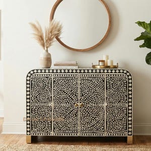 Könnte beinhalten: Ein schwarz-weißer Schrank mit floralem Design. Der Schrank hat vier Türen mit goldfarbener Hardware und steht auf goldenen Beinen. Über dem Schrank hängt ein runder Holzspiegel, und eine Vase mit Trockenblumen steht darauf.