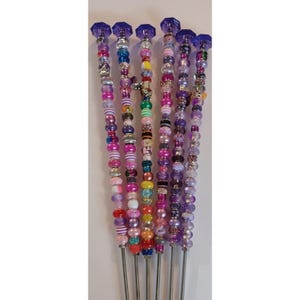 May include: A collection of seven beaded skewers with a variety of colorful beads in shades of pink, purple, and silver. Each skewer has a silver metal stem and a faceted purple crystal top. The beads are arranged in unique patterns.