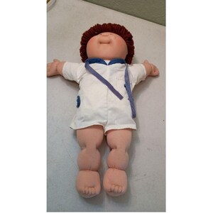 May include: A vintage Cabbage Patch Kid doll with a round face, brown yarn hair, and a white outfit with blue trim. The doll has outstretched arms and soft, stuffed legs and feet.