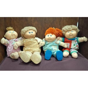 Puede incluir: Cuatro muñecas Cabbage Patch Kids están sentadas una al lado de la otra. Las muñecas tienen caras redondas, ojos azules y varios peinados. Visten diferentes atuendos en colores pastel, incluyendo rosa, amarillo, azul y rojo, con zapatos a juego.