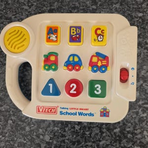 May include: A cream-colored VTech Talking Little Smart School Words electronic learning toy featuring a yellow speaker, three letter buttons with illustrations, three vehicle buttons, and three shape buttons labeled with the numbers one, two, and three.