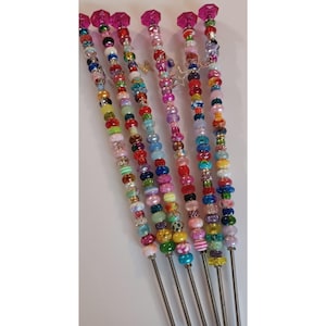 May include: A collection of beaded skewers with a variety of colorful beads in different shapes and sizes. Each skewer has a pink, faceted crystal at the top. The beads are strung on a silver metal rod.