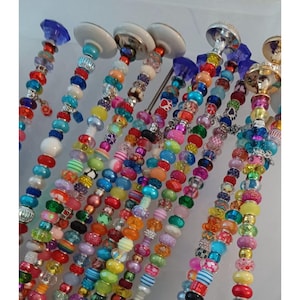May include: A collection of decorative beaded wine bottle stoppers. Each stopper features a unique design with colorful beads in various shapes and sizes. The stoppers have a variety of top designs, including clear, white, and silver-toned metal.